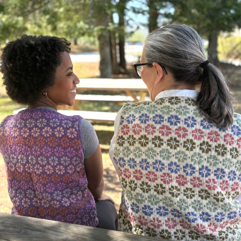 Two women sitting outdoors wearing handknit vests with flowers. Pattern is Flowers of Fortrose Vest.