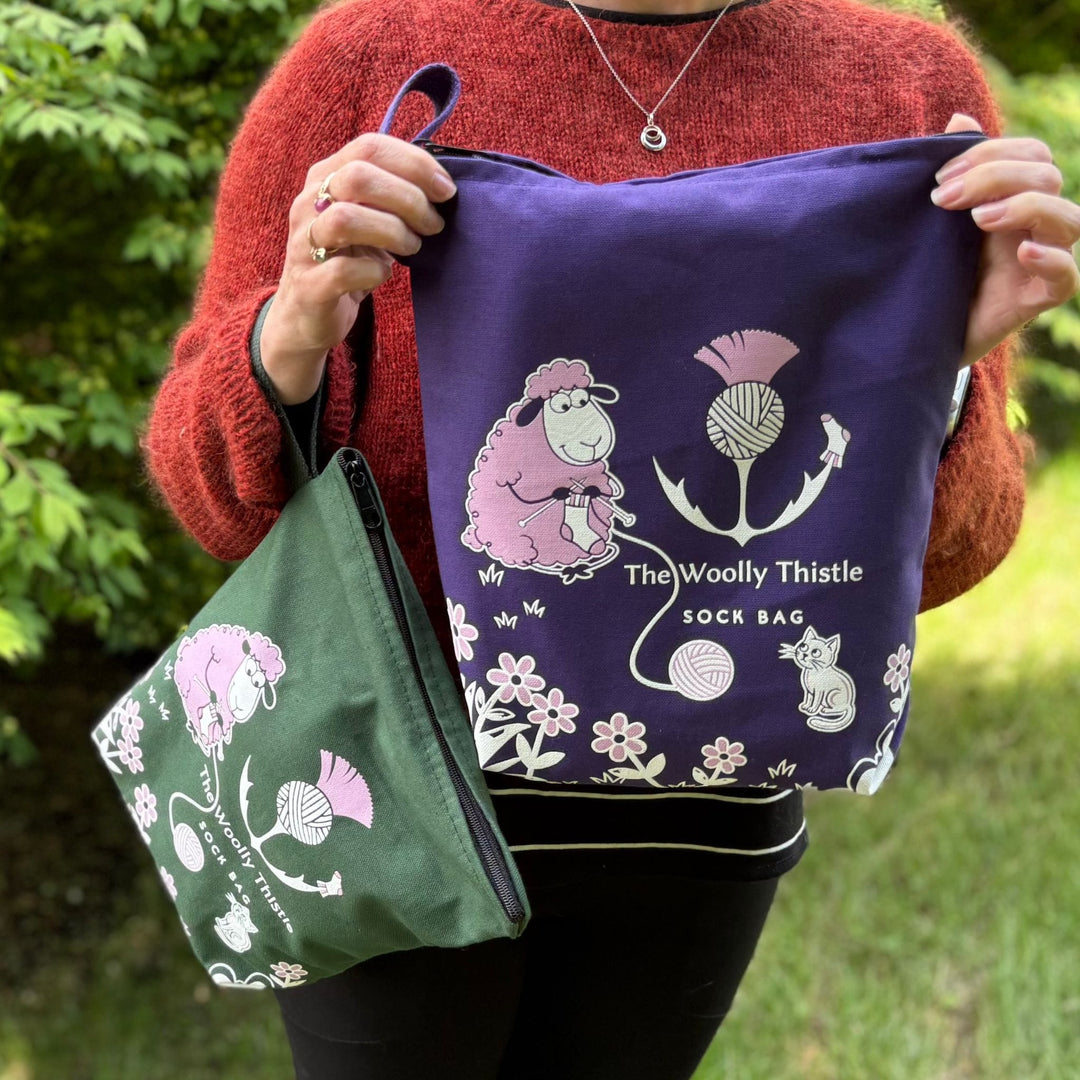 Person holding a purple sock bag outdoors with 'The Woolly Thistle Sock Bag' branding and design on the bag. Bags are in green and purple.