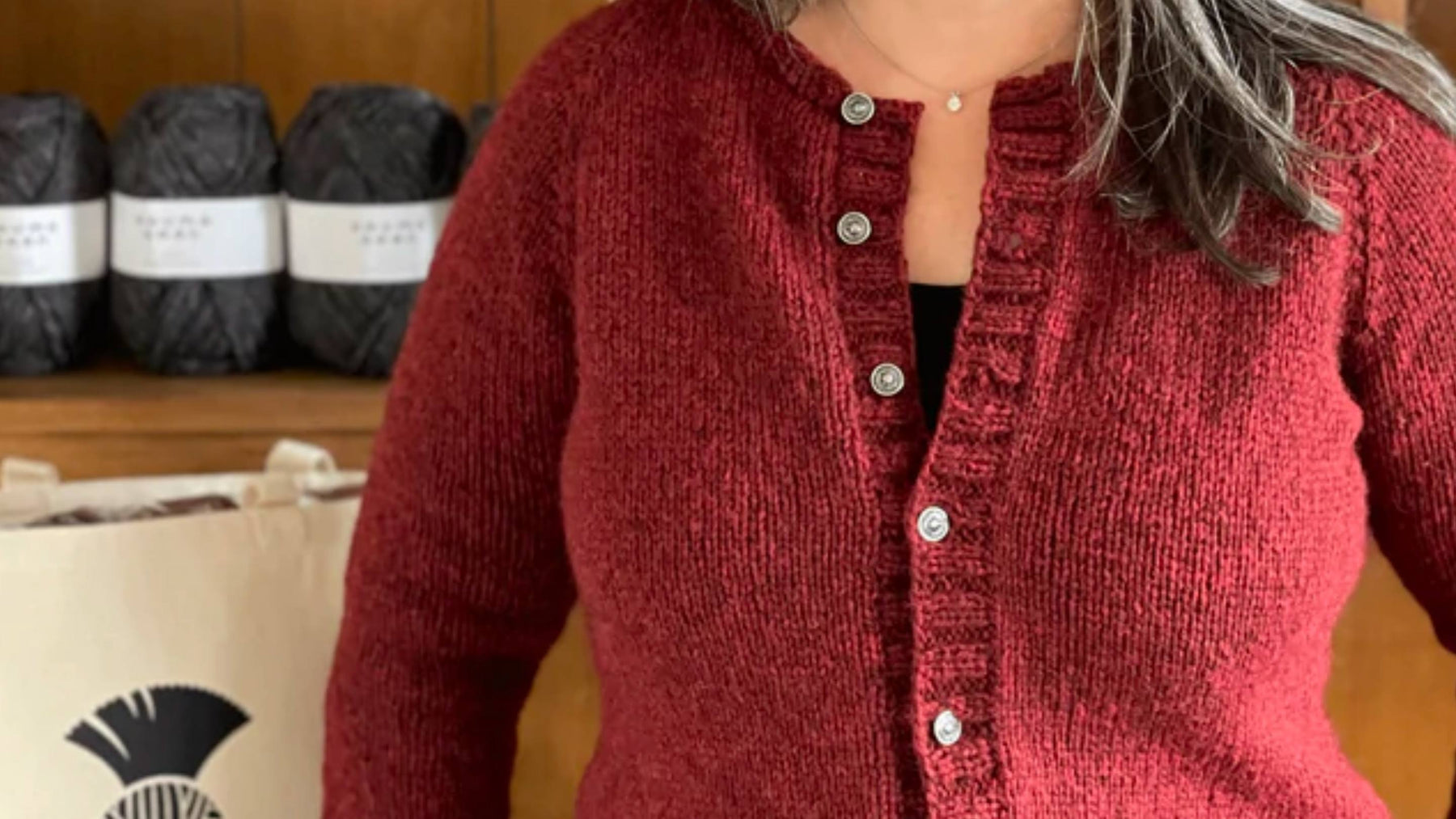 Shoulder and Bust view of woman wearing The Woolly Thistle Victory Cardigan. Sweater is a deep red color with white buttons. Woman is standing in front of a shelf with a TWT Tote bag and Rauma Vams yarn balls. 