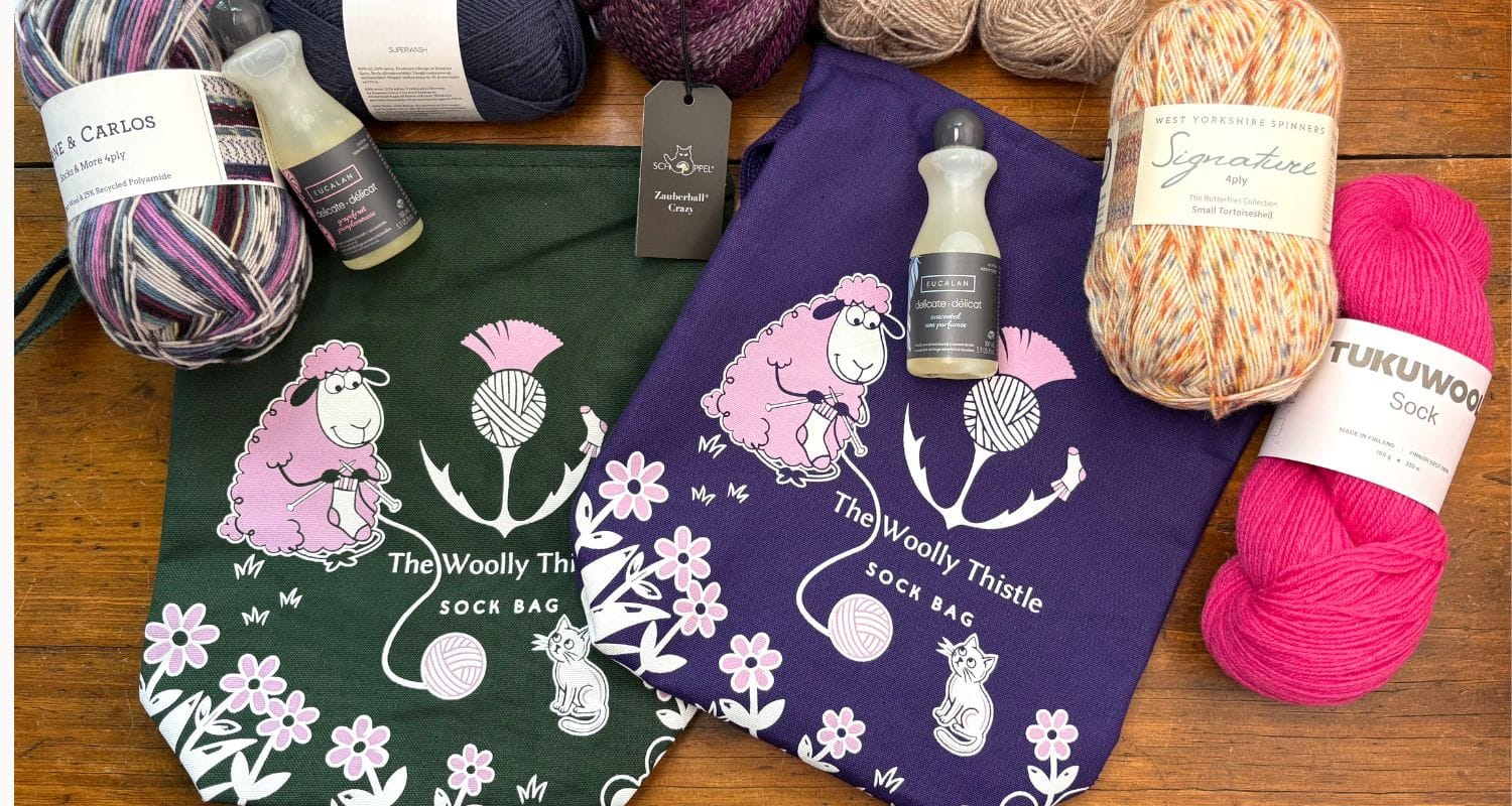 Two bags in green and purple with a knitting sheep. Yarn and knitting supplies surround the bags. Text printed on bag 'The Woolly Thistle Sock Bag'