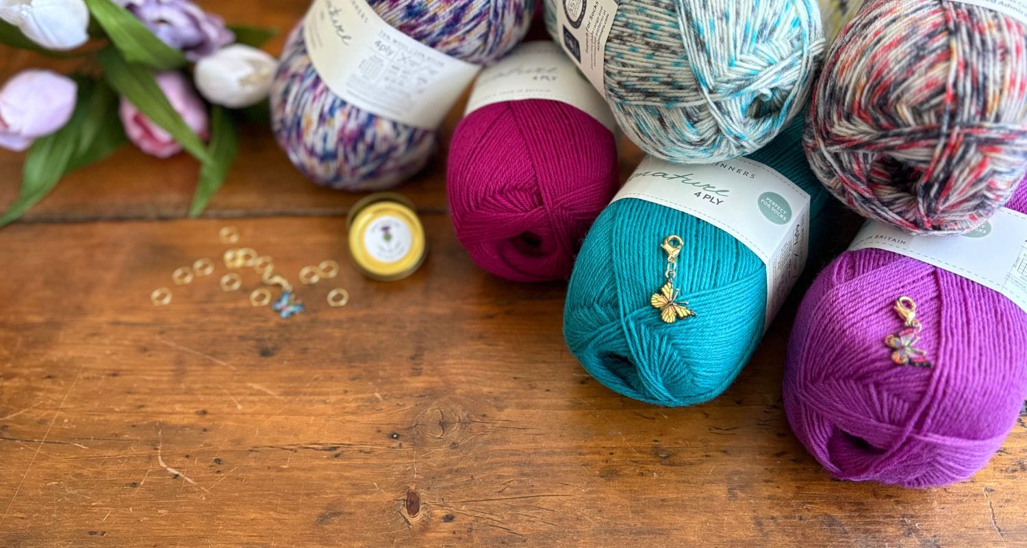 Colorful yarn balls on a wooden surface with flowers and butterfly stitch markers. Yarn is West Yorkshire Spinners Signature 4ply. 