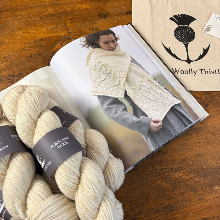 Two skeins of cream-colored yarn with labels on a wooden surface, next to an open book featuring a person wearing a knitted garment.