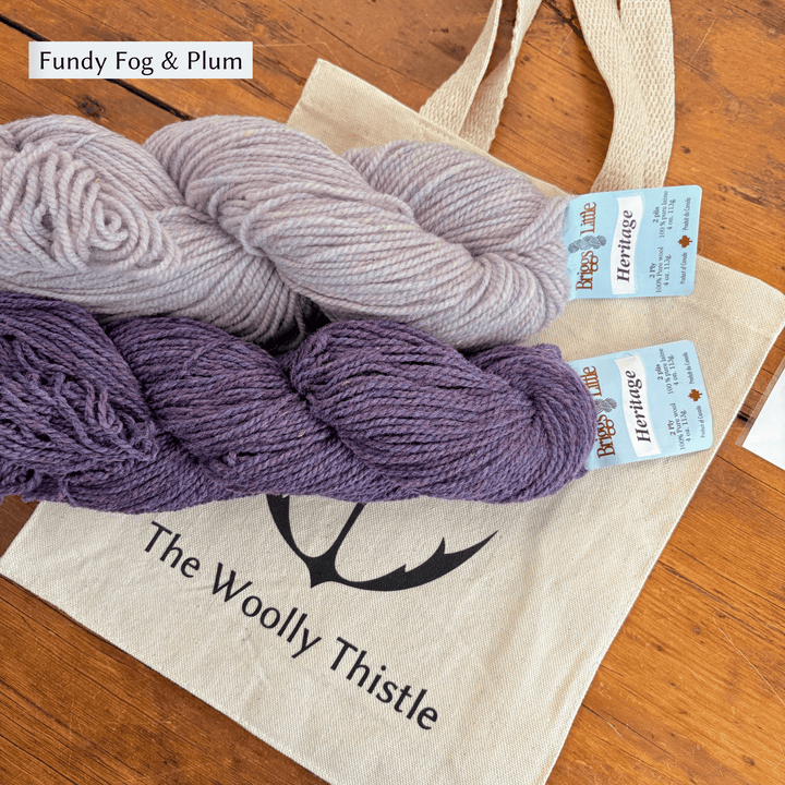 Two skeins of yarn in 'Fundy Fog & Plum' colorway on a tote bag with 'The Woolly Thistle' branding. Yarn set designed to knit Overwinter Mittens and Tam designed by Virginia Sattler-Reimer. 