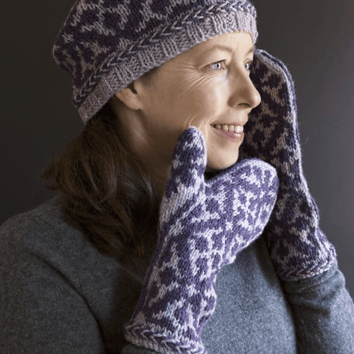 Virginia Sattler-Reimer wearing a matching knitted hat and mittens standing against a dark background. Wearing the Overwinter Tam and Mittens knit in two shades of purple Briggs & Little yarn.  