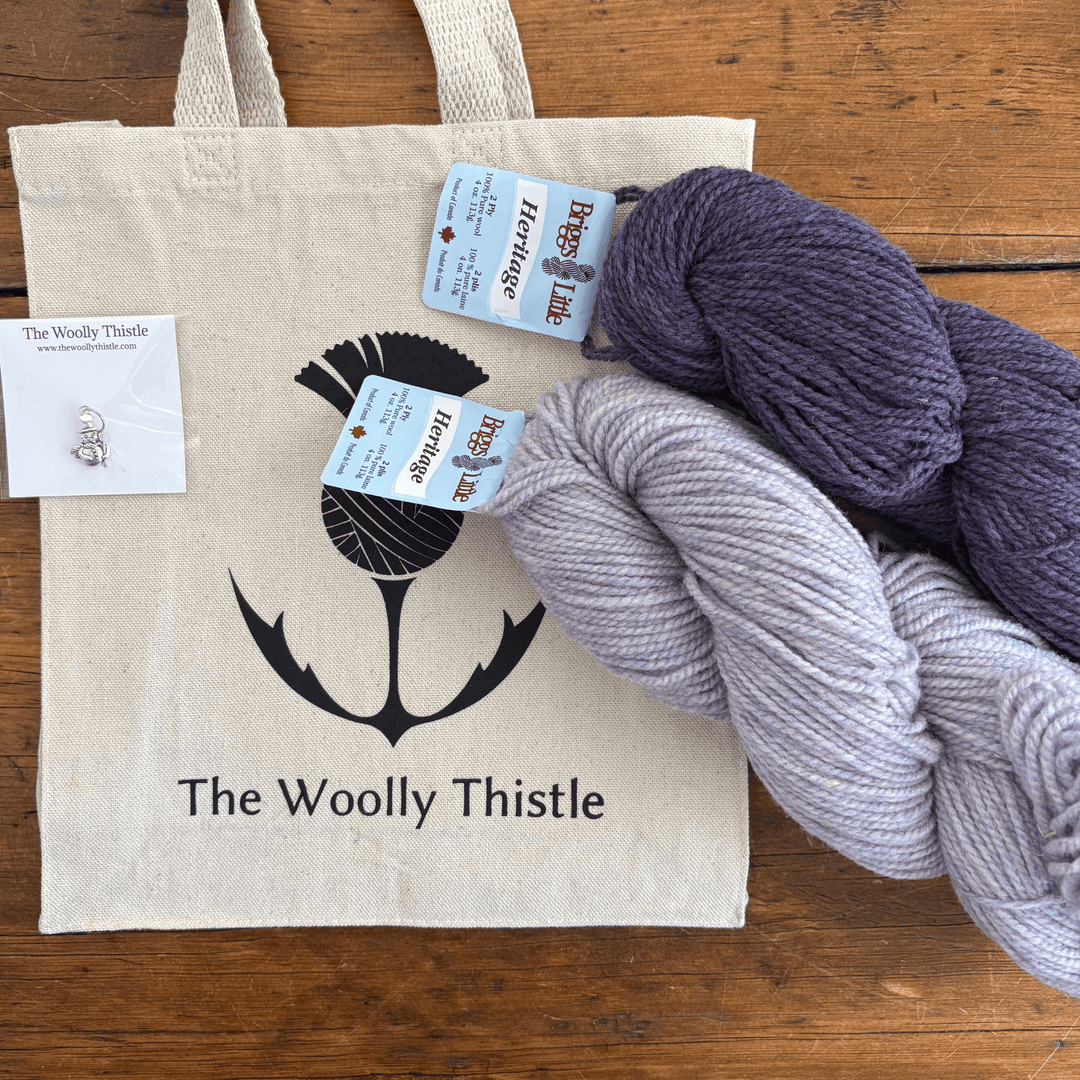 Tote bag with 'The Woolly Thistle' branding, accompanied by skeins of yarn on a wooden surface.Yarn set designed to knit Overwinter Mittens and Tam designed by Virginia Sattler-Reimer. 