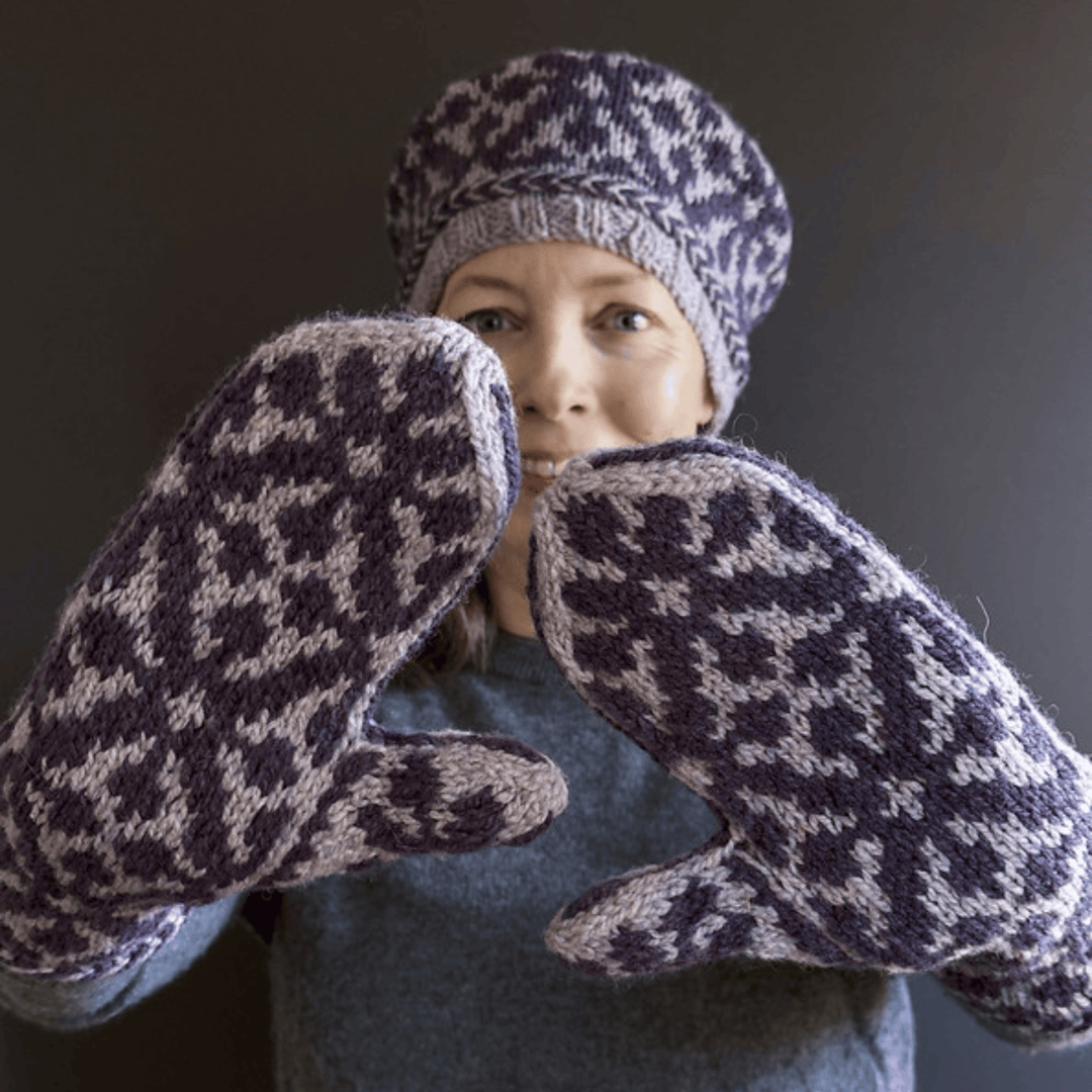 Designer Virginia Sattler-Reimer wearing Overwinter Mittens and Tam knit with Briggs & Little Yarn standing against dark background showing palms of mittens. 
