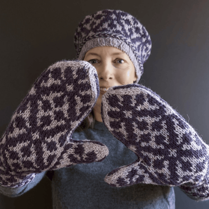 Designer Virginia Sattler-Reimer wearing Overwinter Mittens and Tam knit with Briggs & Little Yarn standing against dark background showing palms of mittens. 