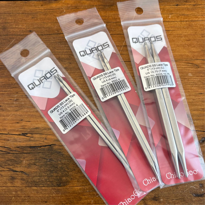 Three packages of Chiaogoo Quads Knitting Needle Tips on a wooden surface 
