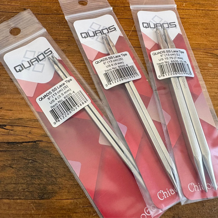 Close up photo of three packages of Chiaogoo Quads Knitting Needle Tips on a wooden surface 