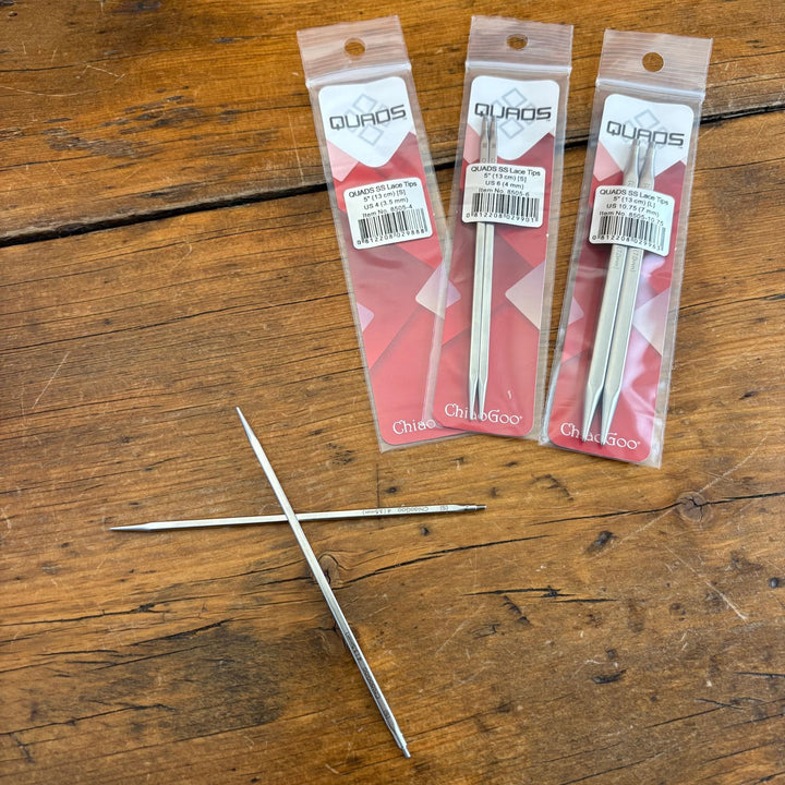 Three packages of Quads needles on a wooden surface. One set of stainless steel needles out of package lying on table. 