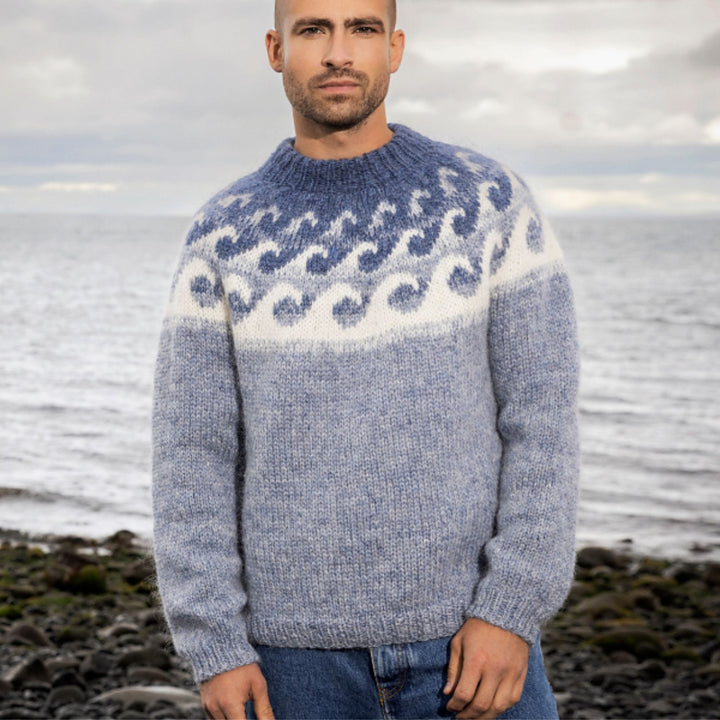 Man wearing a blue patterned sweater called Brimskaflby by Vedis Jonsdotter. He is standing on a beach with ocean in the background