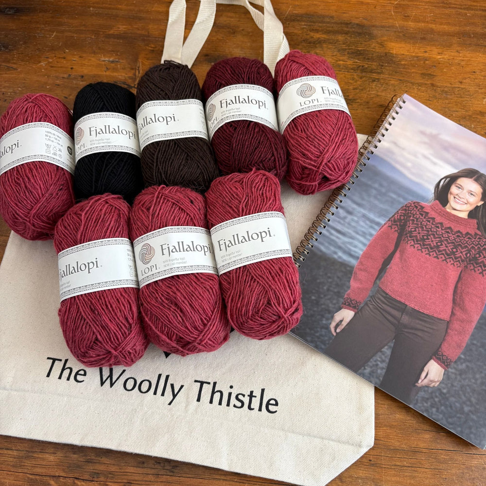 At an angle, Fjallalopi yarn balls in red and brown on a 'The Woolly Thistle' bag with a pattern book showing a person wearing a sweater. Sweater design is the Dalaeti by Vedis Jonsdottir in Fjallalopi.