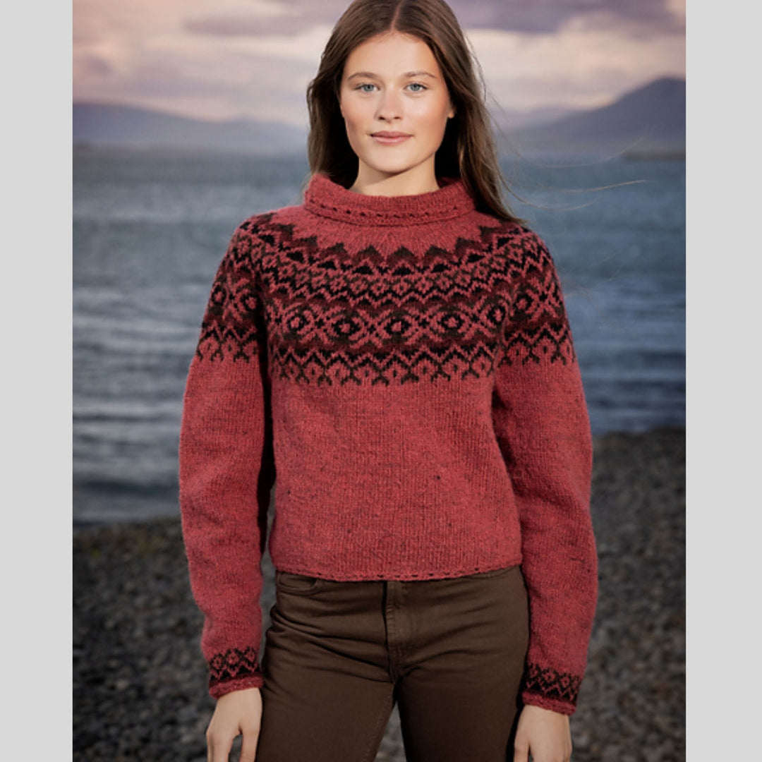 Woman wearing a red sweater with colored yoke standing by a body of water with mountains in the background. Sweater design is Dalaeti by Vedis Jonsdottir in Fjallalopi.