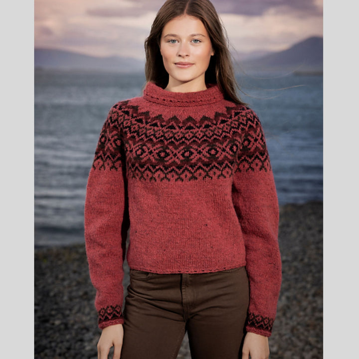 Woman wearing a red sweater with colored yoke standing by a body of water with mountains in the background. Sweater design is Dalaeti by Vedis Jonsdottir in Fjallalopi.