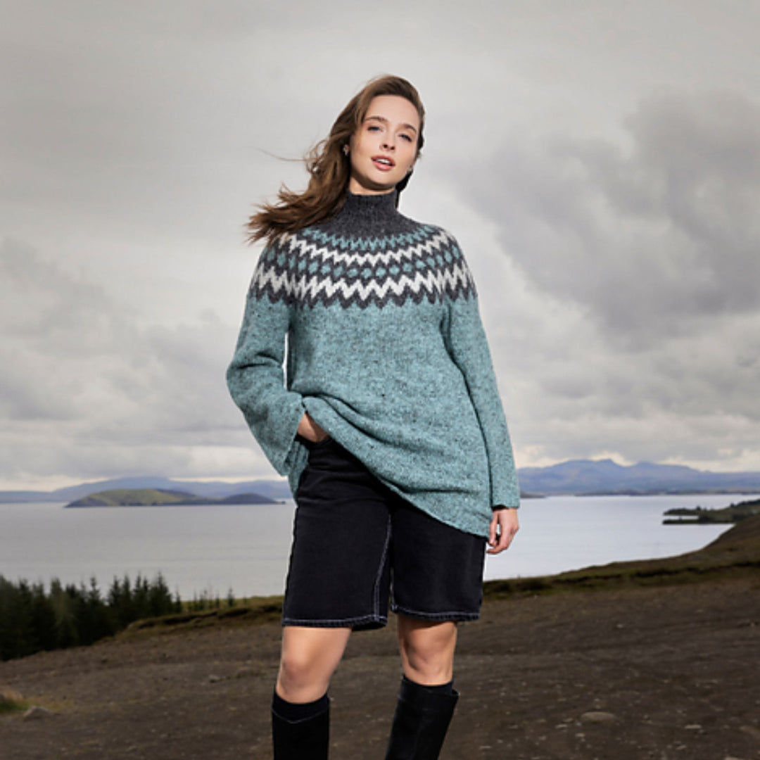 Model wearing the Holt Sweater featuring a teal body and bold Icelandic colorwork yoke, photographed outdoors near water and mountains.