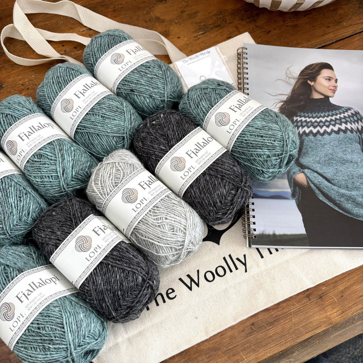 Shown at an angle, Fjallalopi yarn in teal, charcoal, and light gray arranged with the Holt Sweater pattern book, TWT tote bag, and stitch marker.