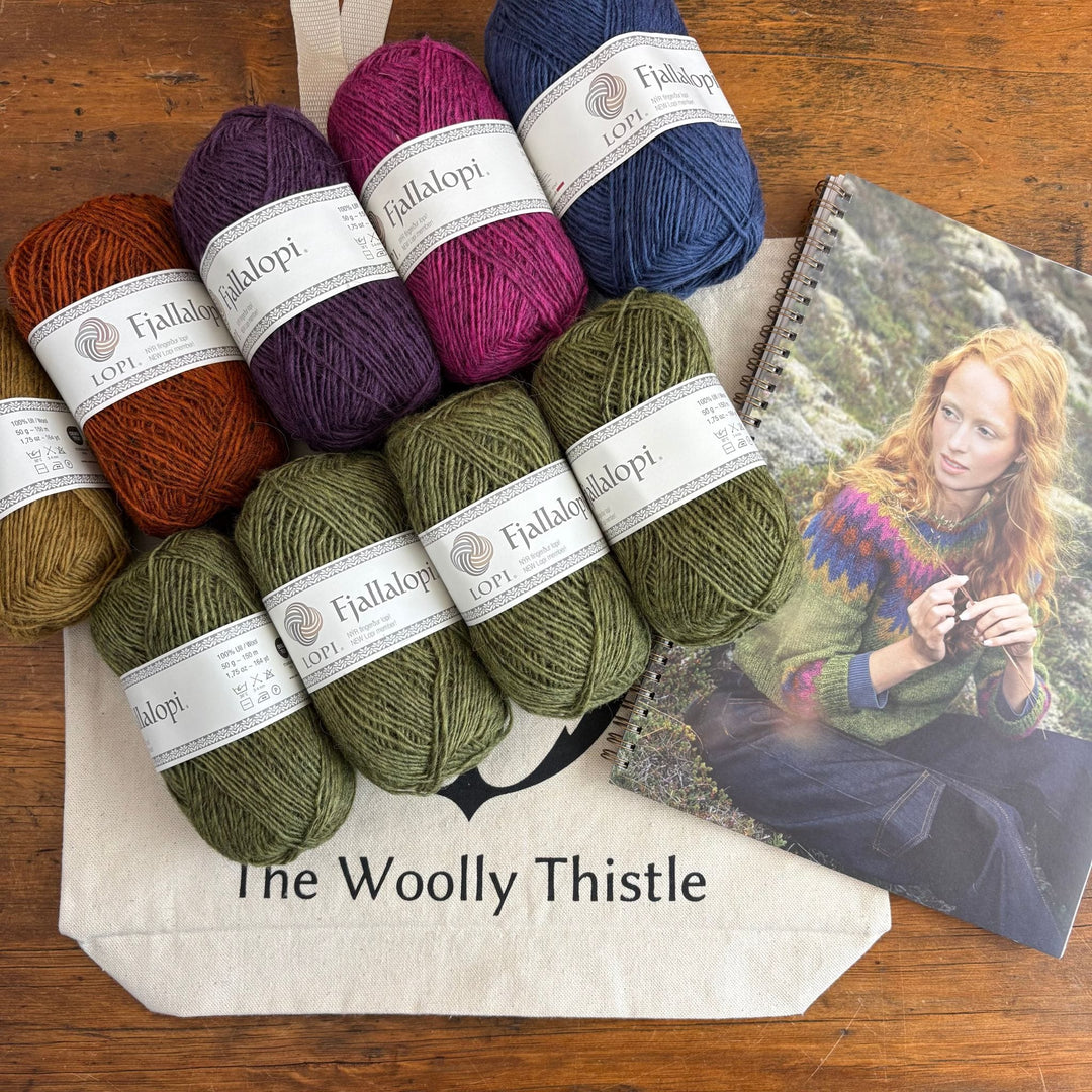Collection of Fjallalopi Yarns with 'The Woolly Thistle' branding tote bag on a wooden surface. Book is open to photo of a colorwork yoke sweater, Mói design which can be knitted with the yarn in the shown.