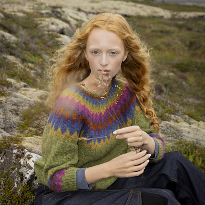 Woman wearing a colorful sweater sitting on a rocky landscape. Sweater is Mói design available in Lopi Book 45. 