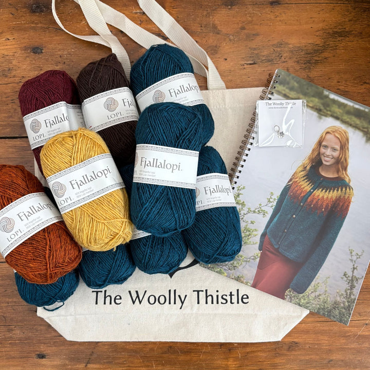 Tigla sweater kit in Fjallalopi yarn including multiple skeins, printed pattern, stitch marker, and The Woolly Thistle tote bag. Flat lay of items on wooden surface.