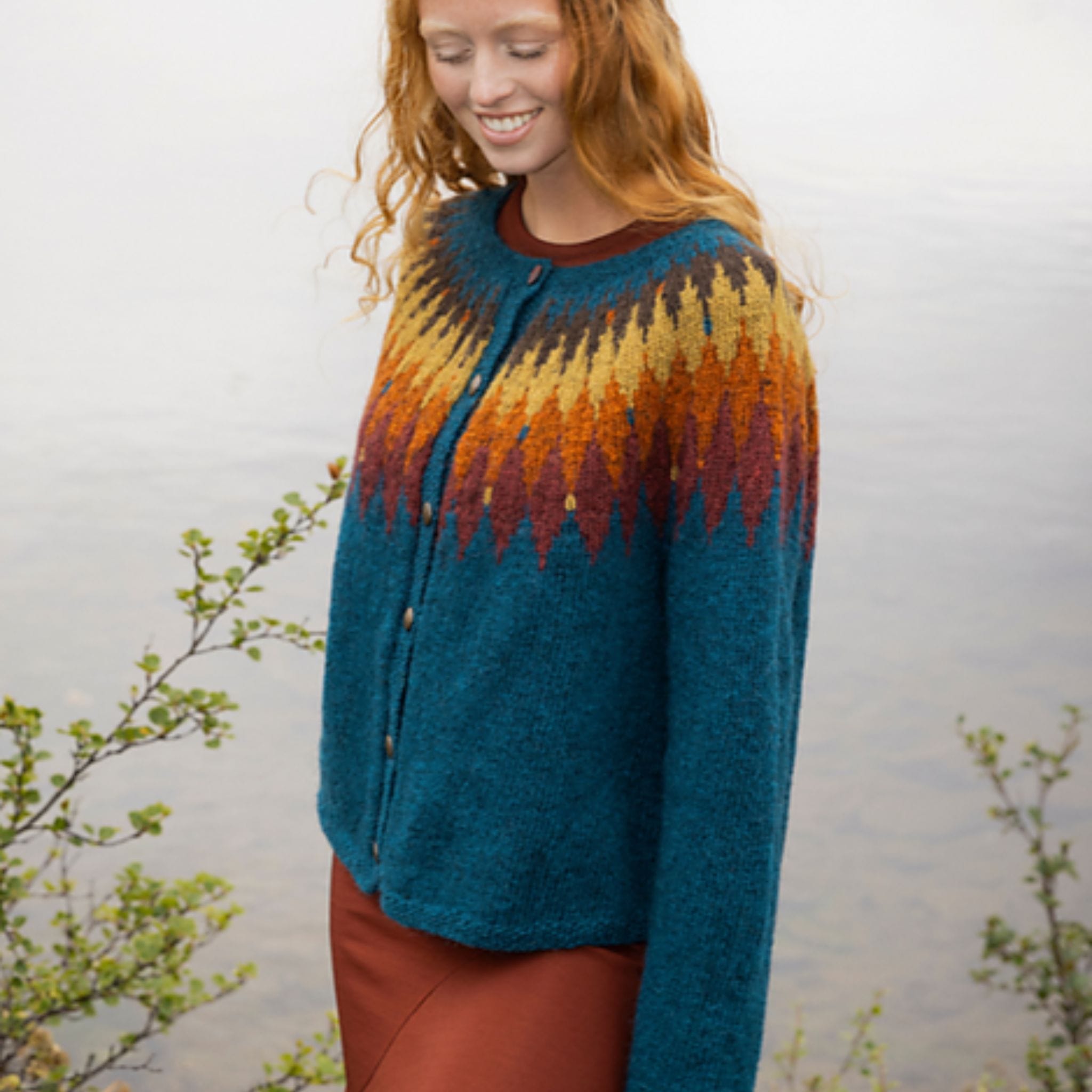 Tigla cardigan by Védís Jónsdóttir knit in Fjallalopi, featuring a teal body with multicolor yoke pattern shown on woman standing sideways in outdoor setting. 