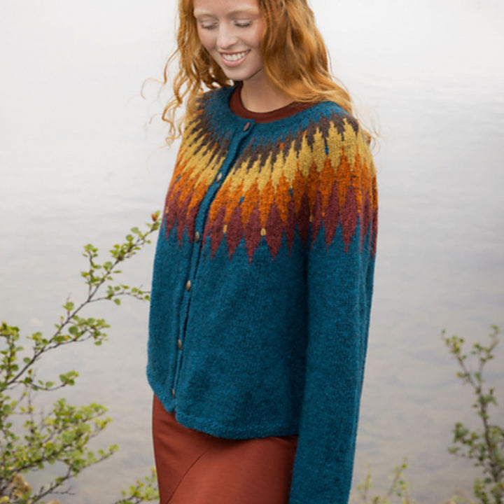 Tigla cardigan by Védís Jónsdóttir knit in Fjallalopi, featuring a teal body with multicolor yoke pattern shown on woman standing sideways in outdoor setting. 