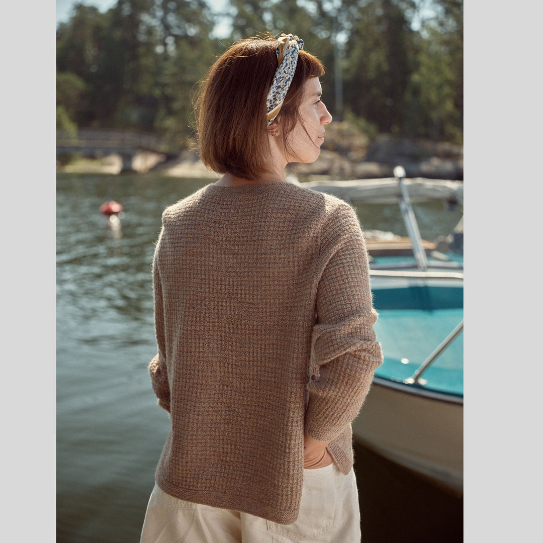 Photo from Laine Issue 28 of back view of a woman wearing beige textured sweater, standing by a lake with boats and trees in the background. 