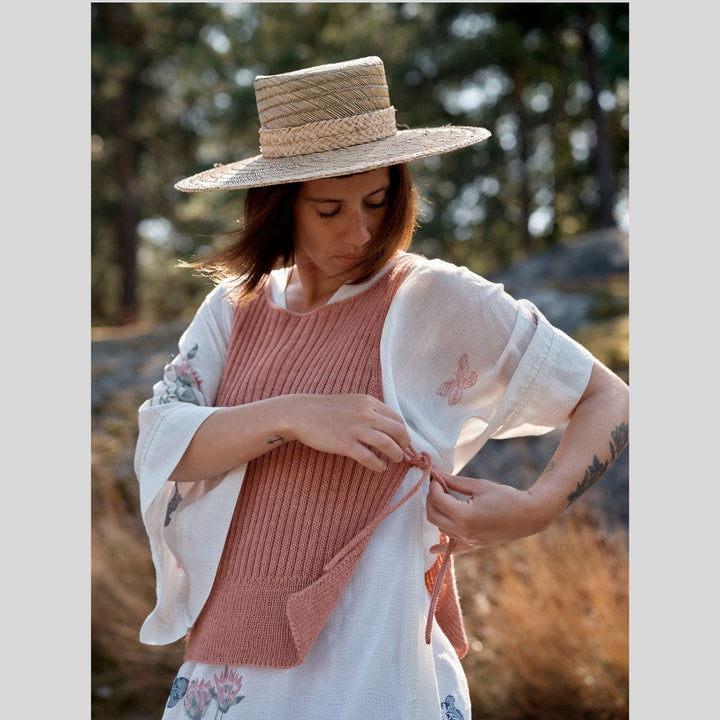 Photo from Laine Issue 28 of woman standing in blurrred outdoor scene, wearing a straw hat and pink vest open at sides. 