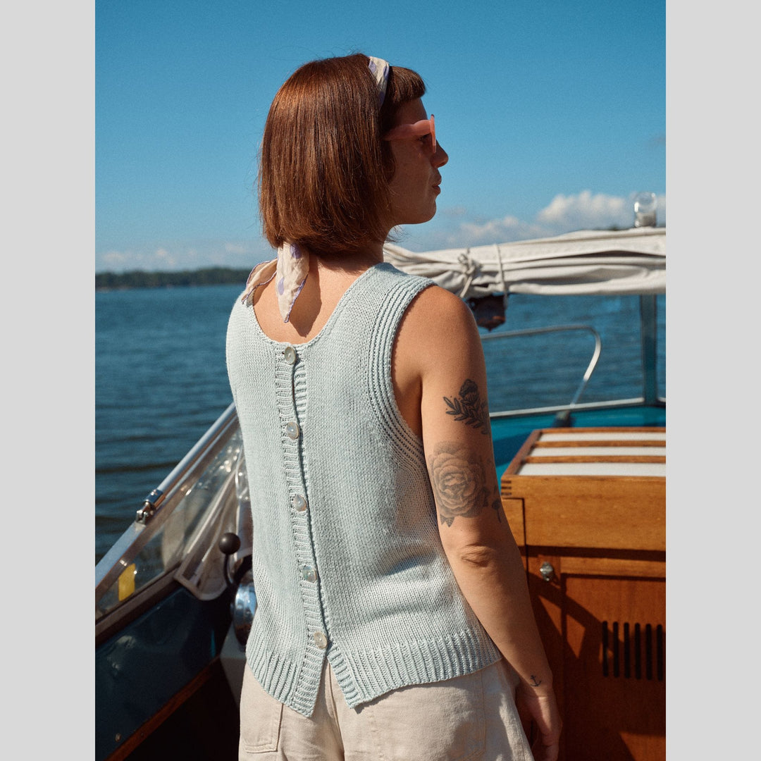 Photo from Laine Issue 28 showing back view of woman on a boat wearing a light blue sleeveless sweater vest with buttons up back with a tattoo visible on her arm.