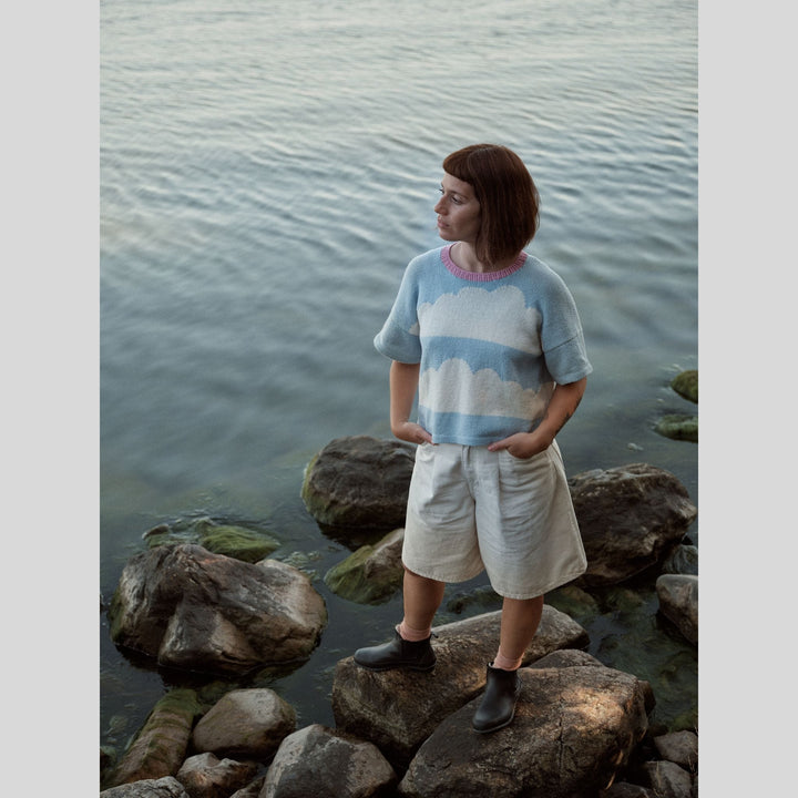 Photo from Laine Issue 28 of a person wearing a cloud-patterned shirt standing on rocks by a body of water.