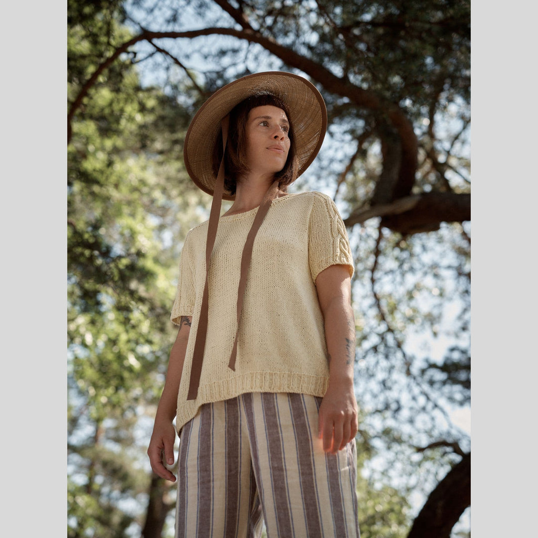 Photo from Laine Issue 28 of a woman wearing a beige/yellow top and striped pants with a wide-brimmed hat, standing among trees.