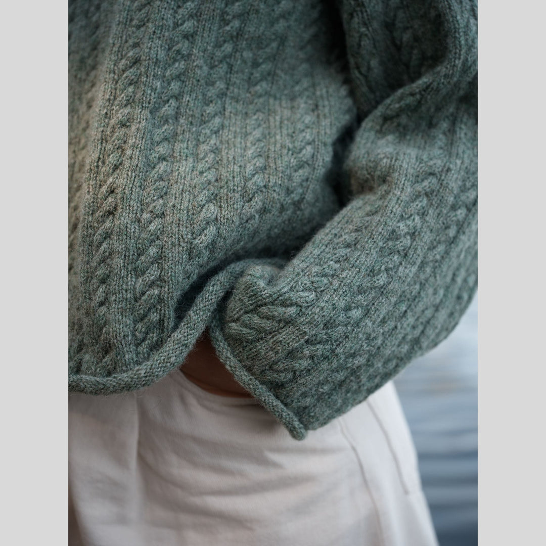 Photo from Laine Issue 28 showing a Close-up of a green knitted sweater arm and body with cable pattern on a blurred background