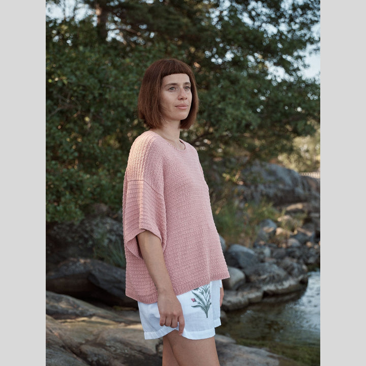 Photo from Laine Issue 28 of a woman wearing a pink sweater and white shorts standing by a body of water with trees in the background