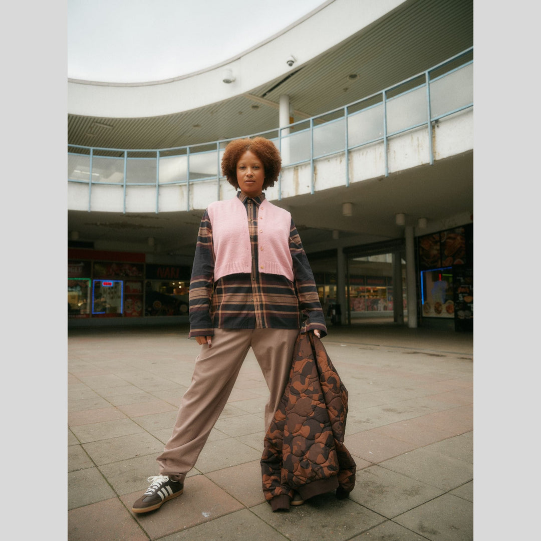 From Laine book My City, My Stitches sold at The Woolly Thistle pages shows a Person wearing a plaid jacket with a pink knit vest in an indoor public space.
