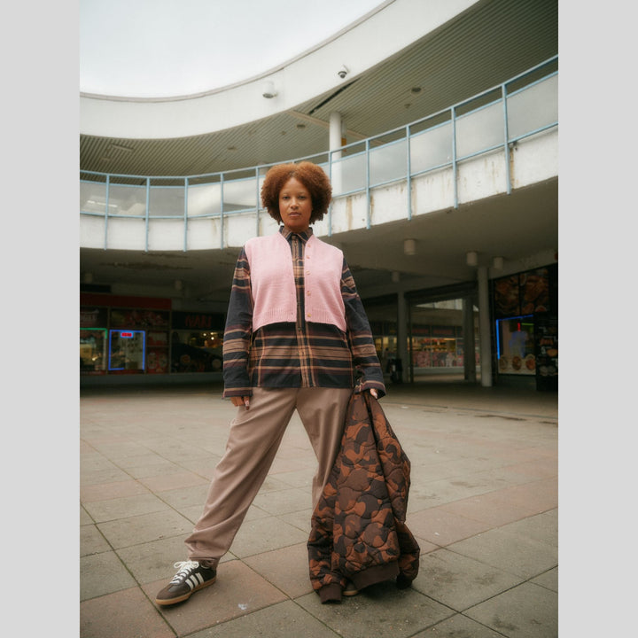 From Laine book My City, My Stitches sold at The Woolly Thistle pages shows a Person wearing a plaid jacket with a pink knit vest in an indoor public space.