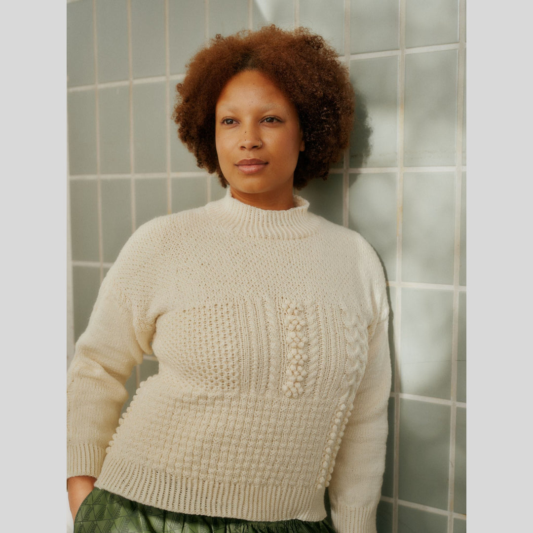 From Laine book My City, My Stitches sold at The Woolly Thistle pages shows a Woman wearing a beige knitted sweater against a tiled wall.