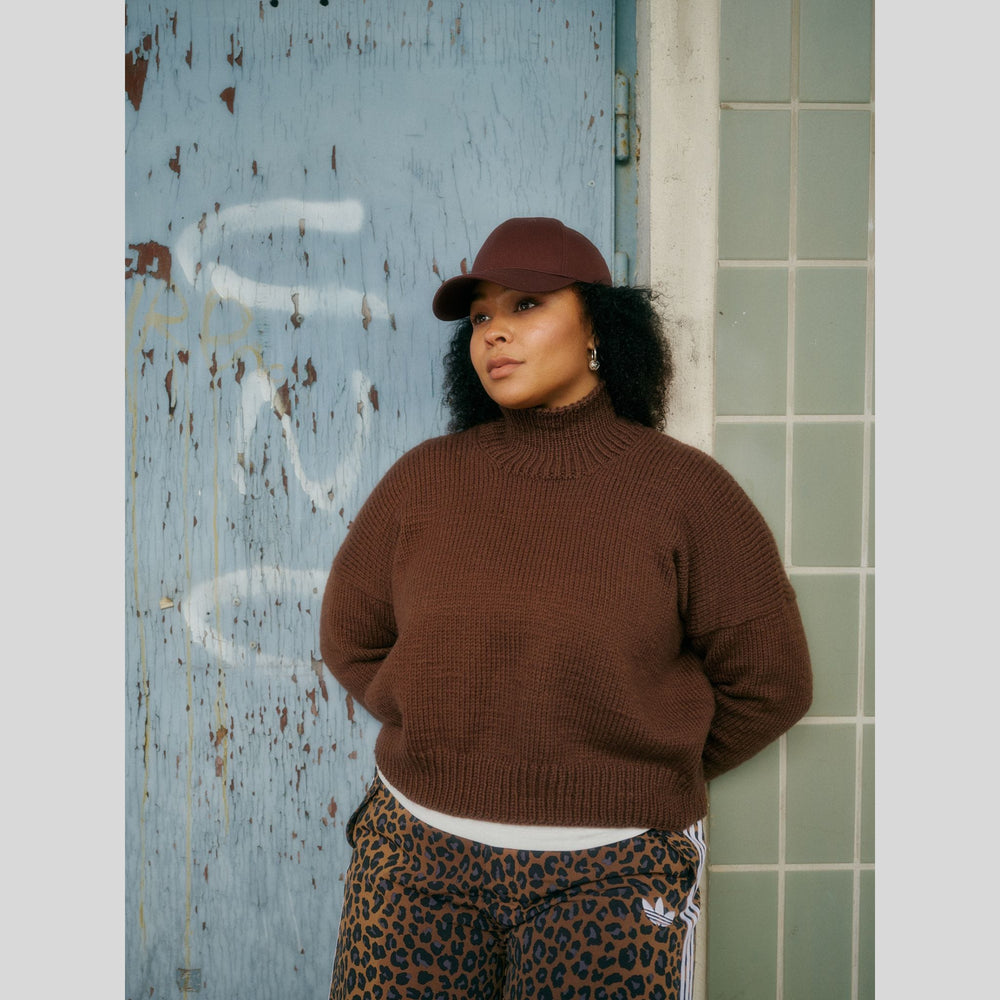 From Laine book My City, My Stitches sold at The Woolly Thistle. Person wearing a brown sweater and leopard print pants standing against a textured blue wall.