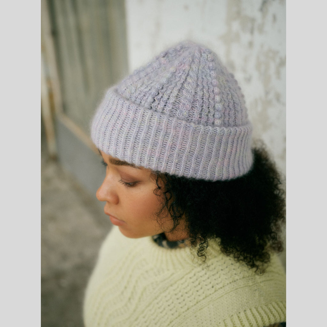 From Laine book My City, My Stitches sold at The Woolly Thistle pages show a Person wearing a gray knit beanie with cables and  a light-colored sweater against a neutral background