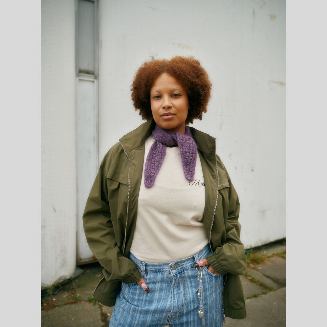 From Laine book My City, My Stitches sold at The Woolly Thistle pages show a Person wearing a green jacket, white shirt, and blue jeans with a purple scarf, standing against a white wall.