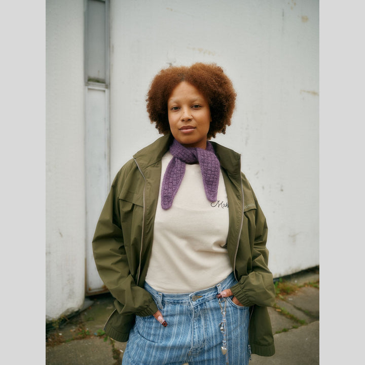 From Laine book My City, My Stitches sold at The Woolly Thistle pages show a Person wearing a green jacket, white shirt, and blue jeans with a purple scarf, standing against a white wall.