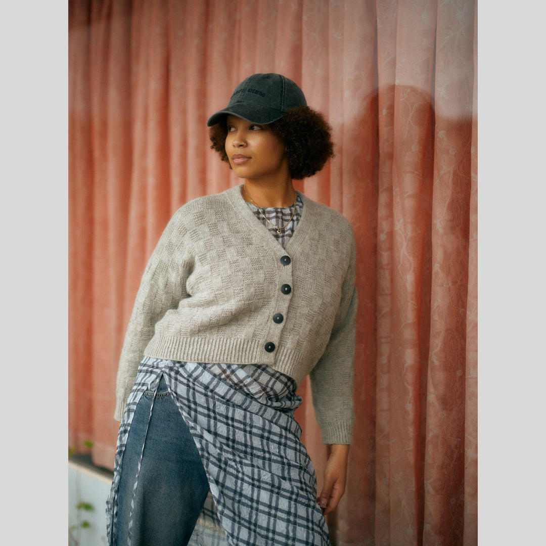 From Laine book My City, My Stitches sold at The Woolly Thistle pages show woman wearing a beige, textured cardigan and leaning against an orange curtain. 