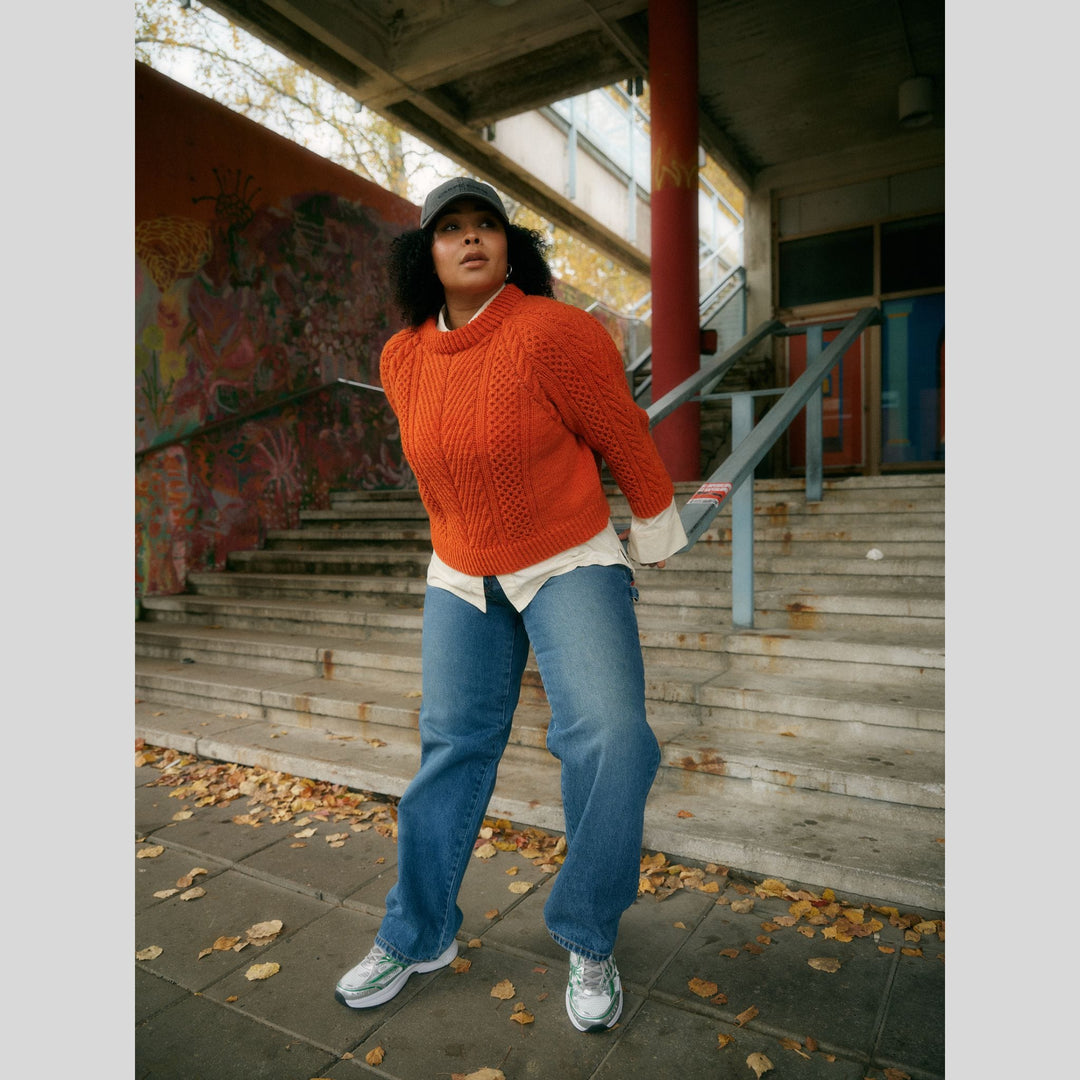 From Laine book My City, My Stitches sold at The Woolly Thistle pages show person wearing an orange, knit sweater with texture and blue jeans standing on a staircase with graffiti in the background.