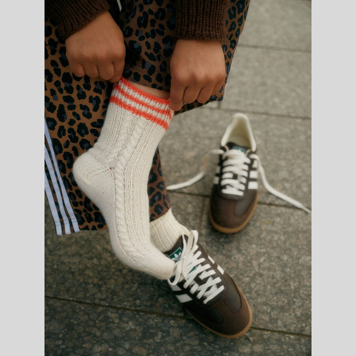 From Laine book My City, My Stitches sold at The Woolly Thistle pages shows person wearing sneakers and showing off handknit socks with orange at top and cable down the side.