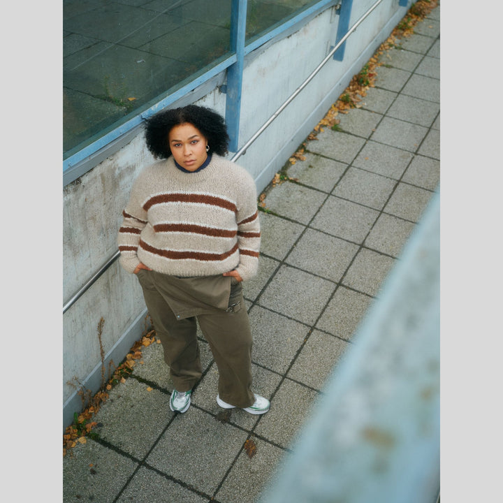 From Laine book My City, My Stitches sold at The Woolly Thistle page shows person wearing a striped sweater and brown pants standing on a sidewalk.