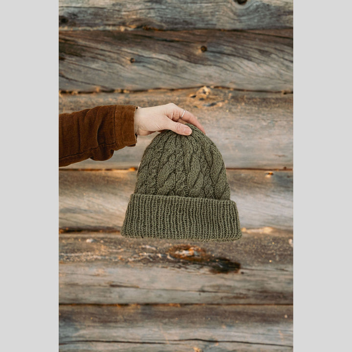 Hand holding a green knitted beanie against a wooden background. This is a page from Laine Book "Unfolding Landscapes"