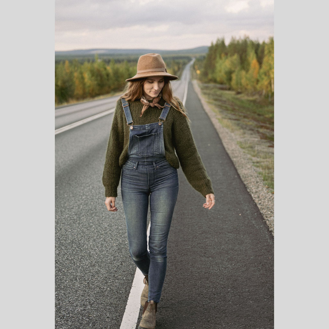 Woman walking on a road with overalls and a hat, surrounded by nature. This is a page from Laine Book "Unfolding Landscapes"