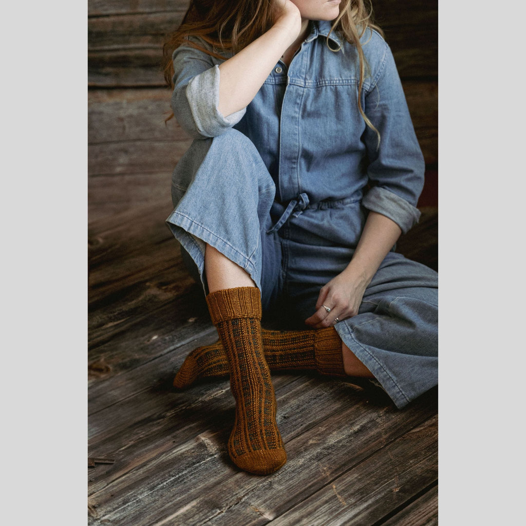 Person wearing a denim dress and brown socks sitting on a wooden floor. This is a page from Laine Book "Unfolding Landscapes"