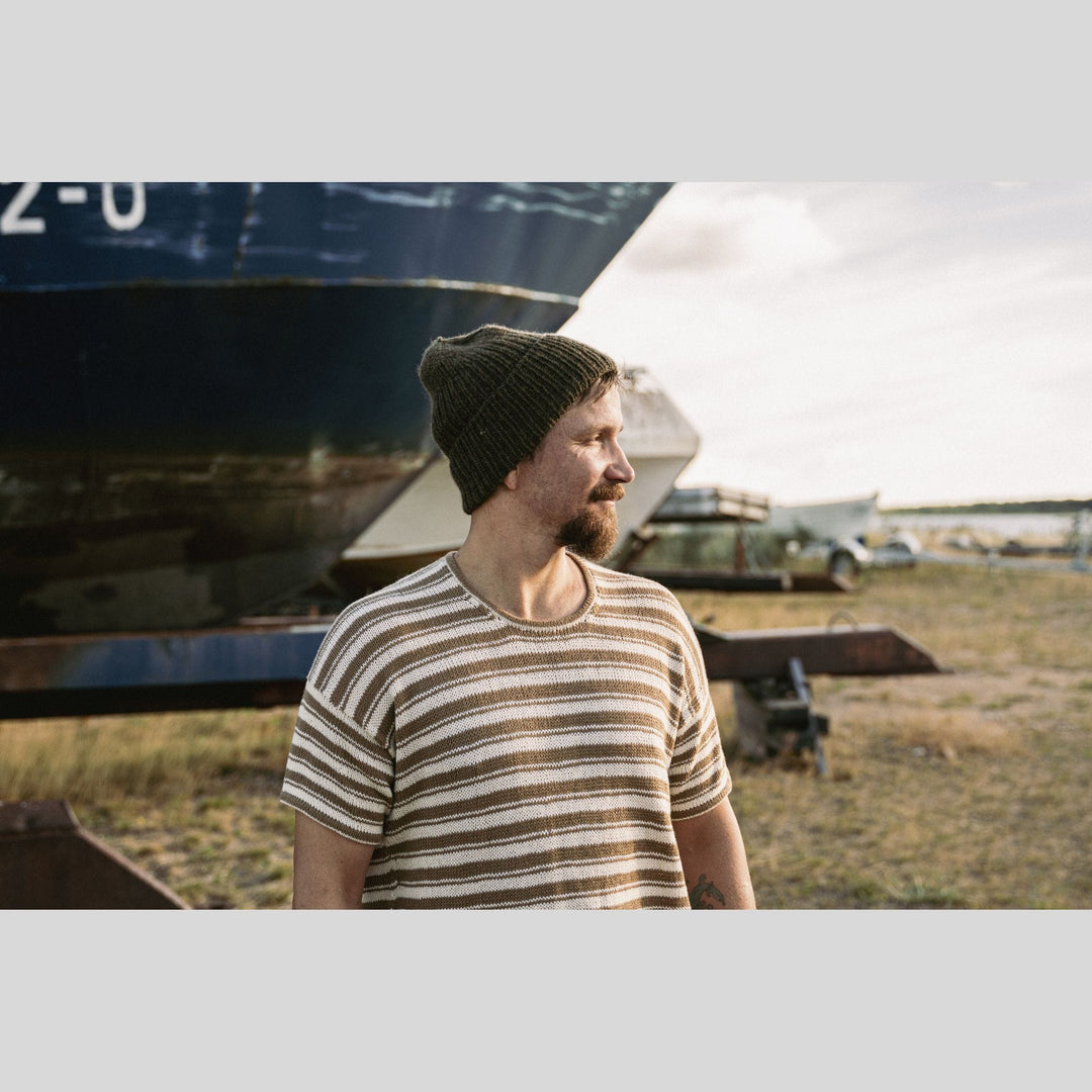 Man wearing a striped  knit shirt and beanie standing in front of boats on a grassy field. This is a page from Laine Book "Unfolding Landscapes"