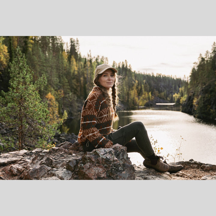Woman sitting on a rock by a lake surrounded by trees. She is wearing a knit sweater. This is a page from Laine Book "Unfolding Landscapes"