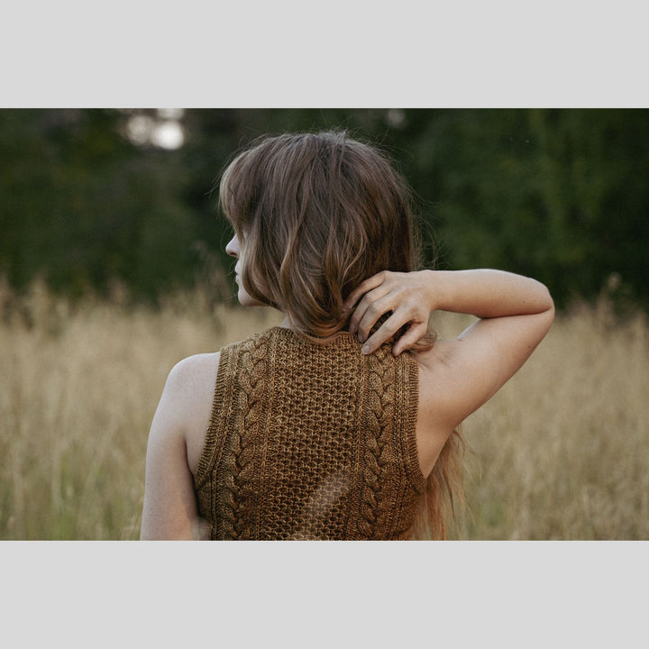 Back view of woman wearing a brown knitted sleeveless sweater in a field. This is a page from Laine Book "Unfolding Landscapes"
