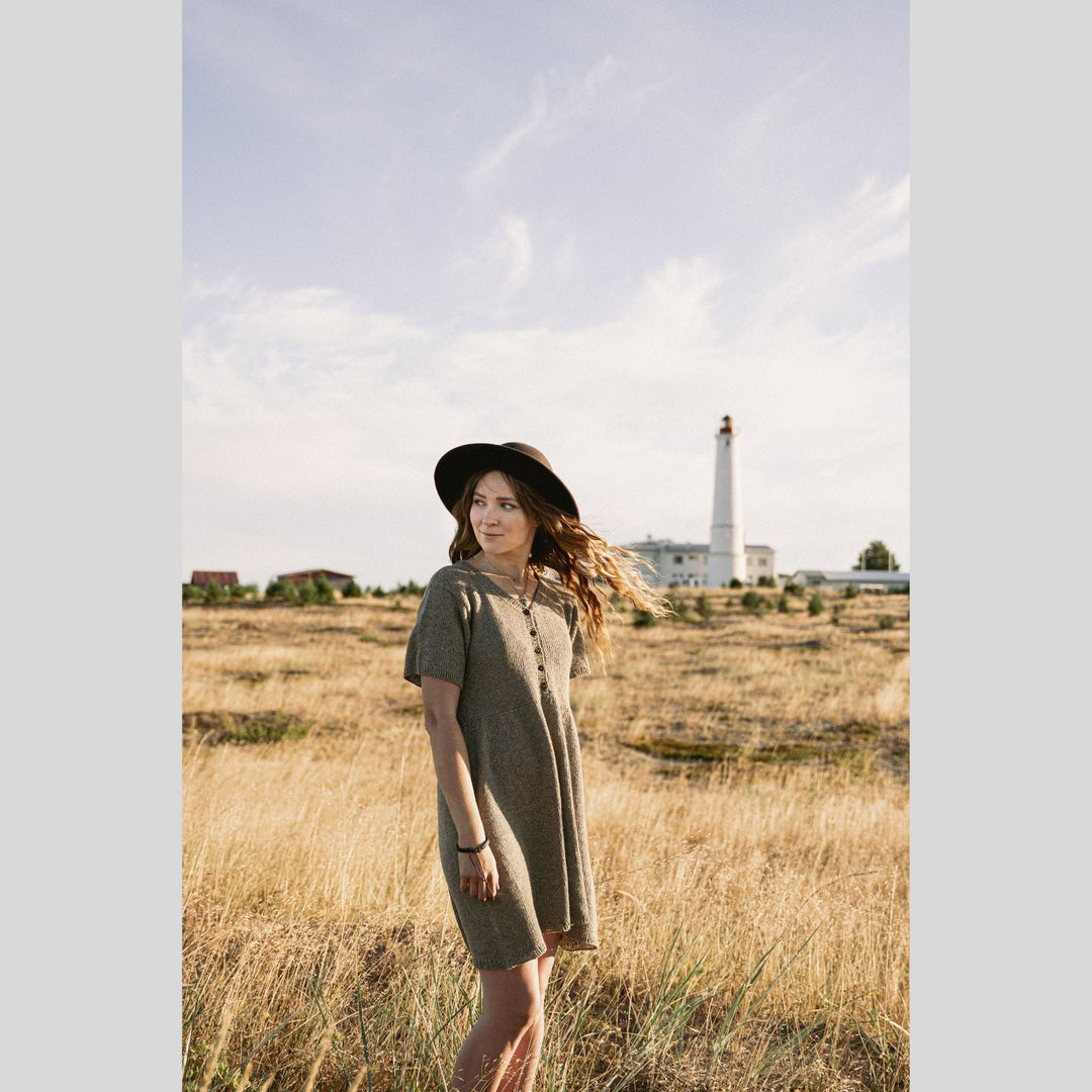 Woman in a knitted dress standing in a field with a lighthouse in the background. This is a page from Laine Book "Unfolding Landscapes"